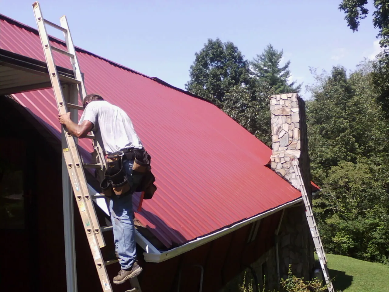 Roofer on a ladder beside a red metal roof and stone chimney — Soffit & Fascia Repair by Roofing Brothers in Haydenville